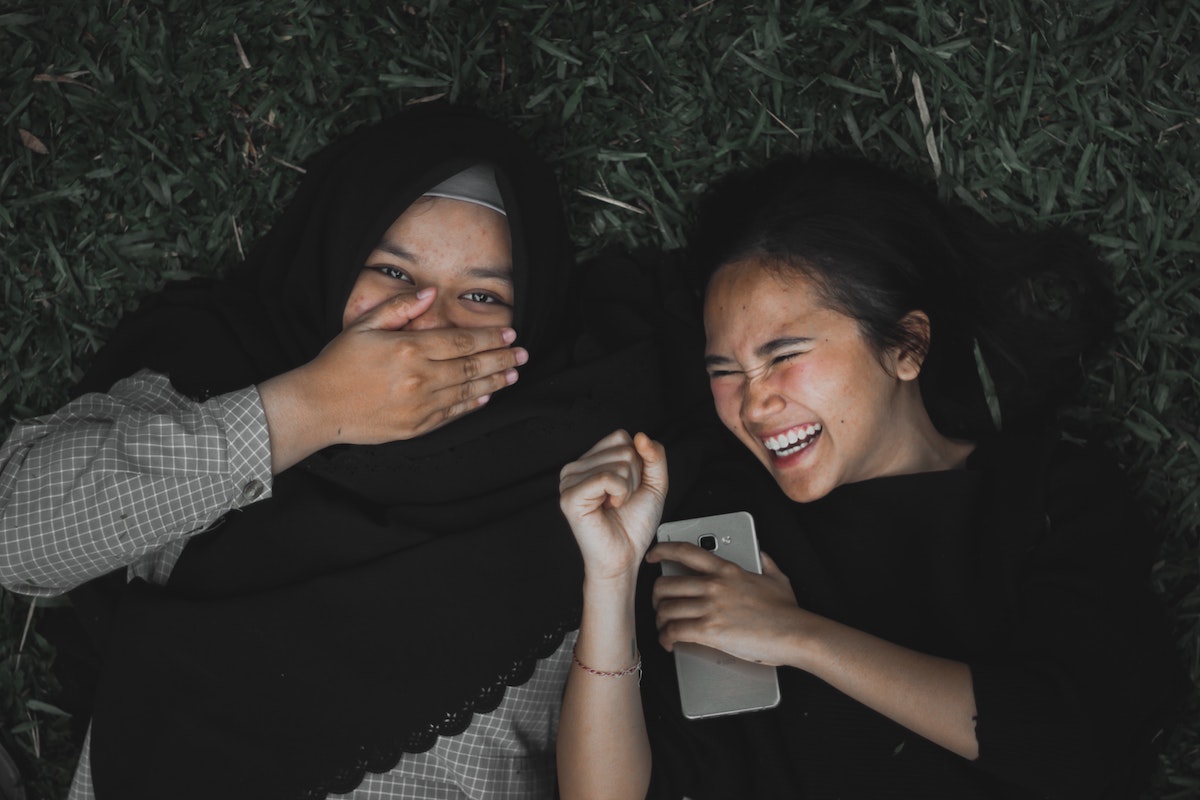Zwei Mädchen lachend auf der Wiese, eine trägt einen Hijab
